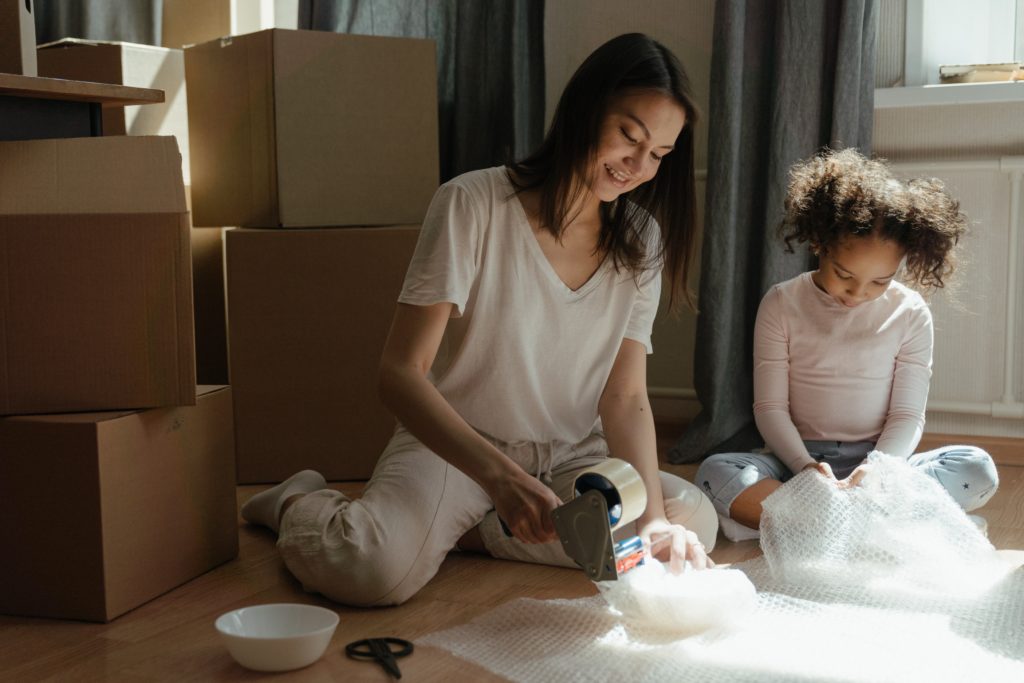Young woman sitting on floor with her young daughter; packing up boxes as they prepare to move.